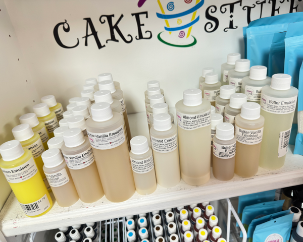 A shelf display of various cake emulsion bottles in different colors (yellow, beige, brown, clear) alongside blue bags of ingredients, with a 'CAKE STUFF' sign on the wall behind them.